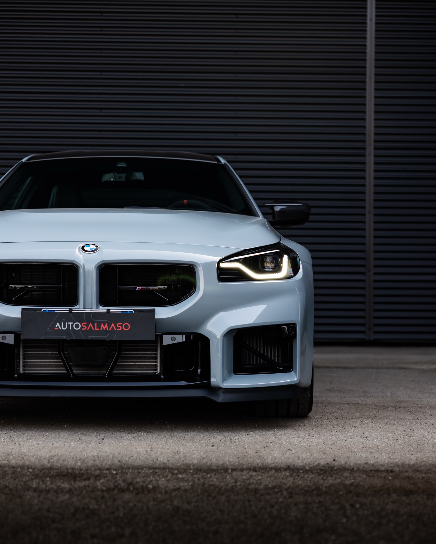 Frontale di BMW M2 grigio chiaro con fari LED accesi e targa AUTOSALMASO, davanti a una saracinesca scura